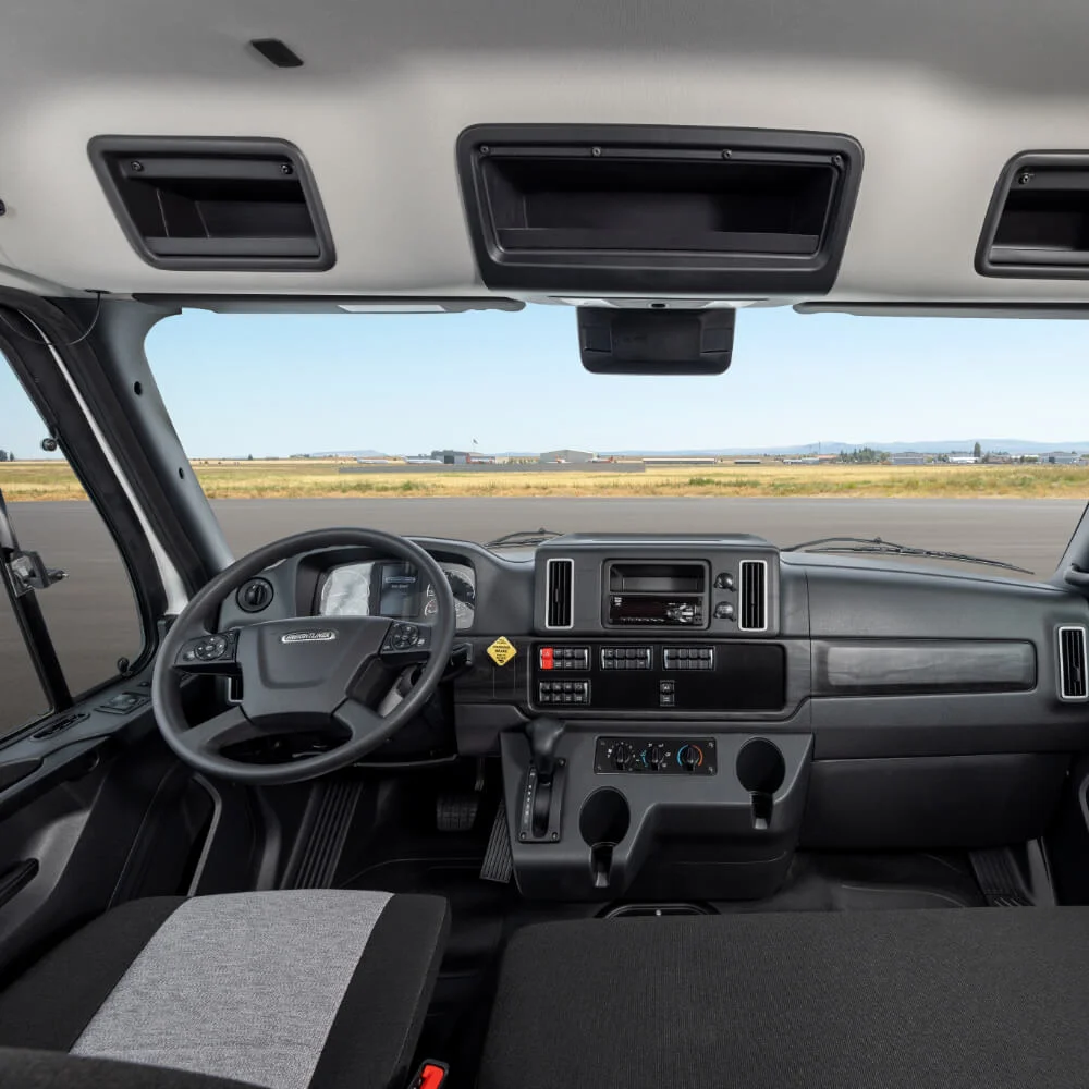 Interior view of the Freightliner 108SD Plus, showcasing a spacious driver-centric design. The ergonomic dashboard layout features clearly labeled controls, a digital display, and a user-friendly infotainment system. The comfortable driver's seat, made of premium fabric, complements the neatly organized cabin. Above, there are practical storage compartments, while the large windshield offers a panoramic view of the open road ahead. The design emphasis on functionality and driver comfort reflects the truck's dedication to operational efficiency and safety. An optimal choice for drivers who value reliability and comfort on long hauls.